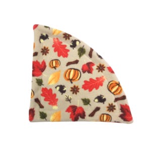 May include: A beige fleece hat featuring a seasonal autumn pattern. The design includes orange pumpkins, red and yellow leaves, brown twigs, and dark brown acorns. The hat is triangular with a rounded top.