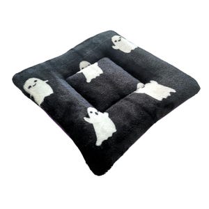 May include: A black and white pet bed with a ghost pattern. The bed is square-shaped with a quilted design and a smaller, raised center cushion. The ghosts are white and have simple, friendly faces.