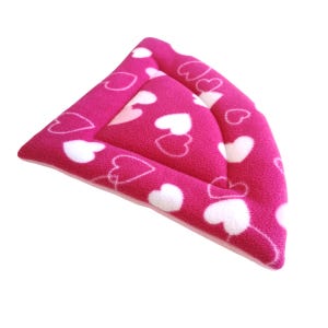 May include: A triangular-shaped pet bed or corner cushion made of soft, bright pink fleece fabric. The surface is covered in a repeating pattern of solid white hearts and outlined heart shapes, featuring a raised border and a padded centre.