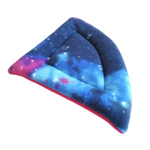 May include: A triangular pet hammock made of soft fleece fabric featuring a vibrant galaxy print with shades of deep blue, purple, and bright pink, accented by white star-like dots. The edges are finished with a solid pink fabric trim.