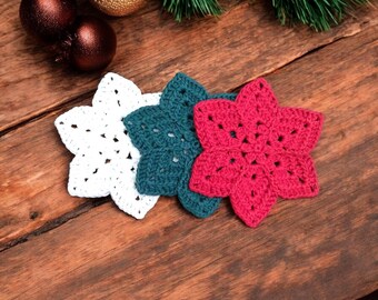 Könnte beinhalten: Drei gehäkelte Weihnachtsstern-Ornamente in Weiß, Türkis und Rot, angeordnet auf einer Holzoberfläche. Die Ornamente sind sternförmig mit durchbrochenen Designs. Weihnachtsdekoration mit Tannenzweigen und Kugeln im Hintergrund.
