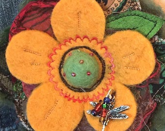 May include: A handmade, colorful flower brooch. The brooch features a large, yellow felt flower with a green center and a dragonfly charm. The flower is attached to a denim jacket, showcasing the brooch's potential use as an accessory.
