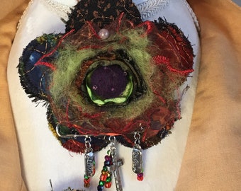 May include: A handmade textile brooch in the shape of a flower, featuring layers of fabric in various colors including red, green, and black. The brooch has dangling charms with the words "love" and "believe", a cross, and colorful beads. It is pinned to a white mannequin.