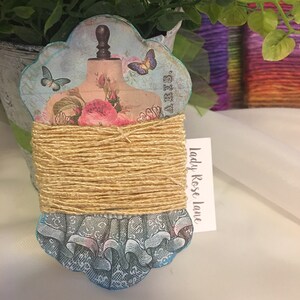 May include: A decorative spool featuring a vintage dress form design with butterflies and roses, wrapped with natural twine. The base has a blue and white lace pattern. The text "Paris" is visible. A tag reads "Lady Rose Lane".