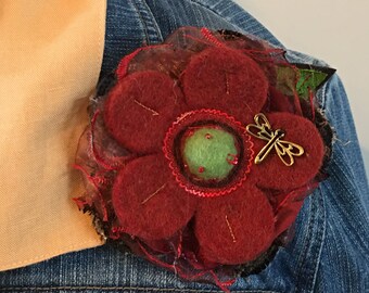 May include: A handcrafted red felt flower brooch with a gold dragonfly accent. The flower has a green center and is attached to a denim jacket. The brooch is a unique accessory.