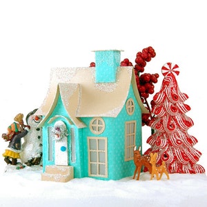 Puede incluir: Una casa en miniatura de color azul turquesa con una puerta y ventanas blancas, rodeada de nieve y decorada con un muñeco de nieve, dos ciervos y un árbol de Navidad de bastón de caramelo.