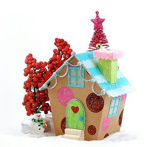 Puede incluir: Un adorno de casa de jengibre caprichoso con un patrón de lunares rosa y rojo, una puerta verde con un corazón y un techo azul y blanco. La casa está decorada con purpurina blanca y un pequeño muñeco de nieve se encuentra frente a ella.