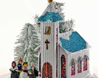 Op de afbeelding: Een witte kerk met een blauw dak en glas-in-loodramen, omringd door met sneeuw bedekte bomen. Vier figuren staan voor de kerk, waaronder een sneeuwpop.
