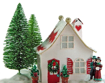 Op de afbeelding: Een wit papieren huis met een rood dak en een rode deur, versierd met een rood hart en een krans. Twee figuren staan voor het huis, een houdt een boek vast en de ander een muziekinstrument. Het huis is omringd door sneeuw en twee kleine groene bomen.