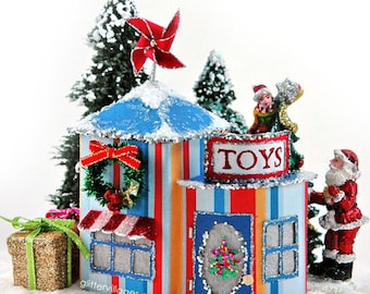Op de afbeelding: Een miniatuur speelgoedwinkel met een blauw-wit gestreepte buitenkant, een rood-witte luifel en een bord met de tekst "TOYS". Er staat een kleine kerstboom voor de winkel en een kerstmanfiguur ernaast. De winkel is versierd met een krans, een windmolen en sneeuw.