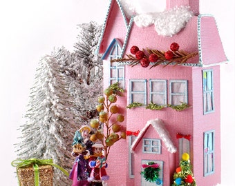 Op de afbeelding: Een roze papieren huis met een besneeuwd dak, versierd met rode bessen, groene krans en een kleine kerstboom. Het huis heeft blauwe versieringen en ramen, en een wit hek. Het huis is omgeven door een besneeuwd landschap met een klein cadeaudoosje en een witte kerstboom.