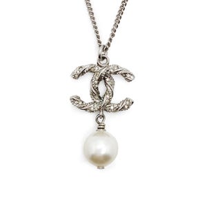 May include: Silver-toned necklace featuring a double-C pendant encrusted with small, clear crystals. A large, round, white pearl dangles below the pendant. The necklace chain is a simple, linked design.