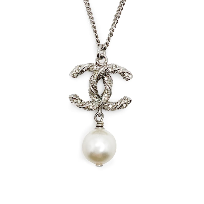 May include: Silver-toned necklace featuring a double-C pendant encrusted with small, clear crystals. A large, round, white pearl dangles below the pendant. The necklace chain is a simple, linked design.
