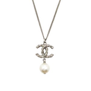 May include: Silver-toned necklace featuring a double-C pendant encrusted with small crystals and a dangling white pearl. The chain is a delicate link style, and the pendant is centered. The background is plain white.