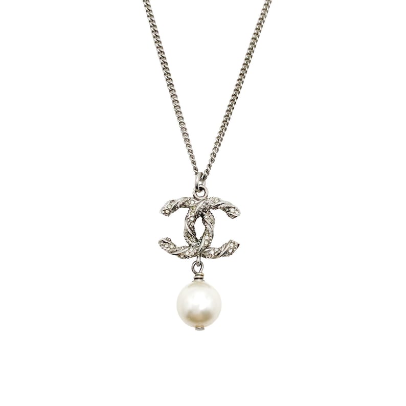 May include: Silver-toned necklace featuring a double-C pendant encrusted with small crystals and a dangling white pearl. The chain is a delicate link style, and the pendant is centered. The background is plain white.