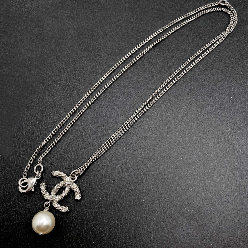 May include: Silver-toned necklace featuring a double-C pendant with a pearl drop. The chain is delicate, and the pendant has a textured design. The necklace is displayed against a dark background.
