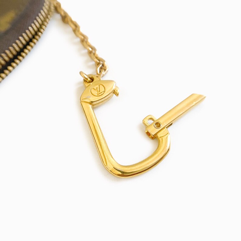 May include: Close-up of a gold-colored metal clasp with a chain and a small, engraved logo. The clasp is open, revealing its hook-like design. The background is white.