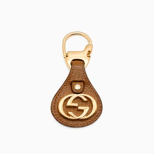 May include: A brown leather keychain with a gold-colored metal clasp and a gold-colored double-G logo. The keychain has a teardrop shape and a small gold-colored button detail.