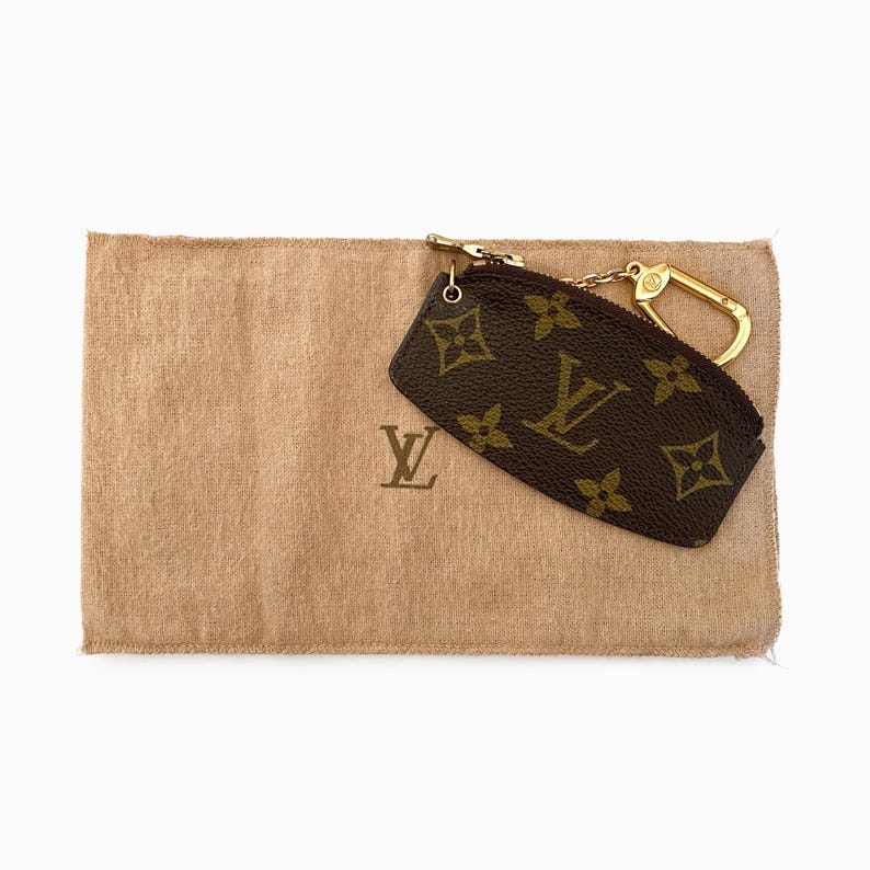 May include: A brown and tan key pouch with a gold-tone clasp and zipper. The pouch features a repeating pattern of brown floral and "LV" logos. It rests on a tan fabric pouch with a matching logo.