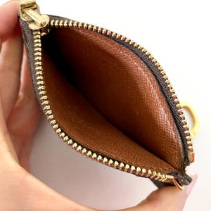 May include: Open brown and tan cardholder with a gold zipper and pull. The interior is a lighter brown with a textured pattern. The zipper is gold-toned, and the cardholder has a small gold ring attached.