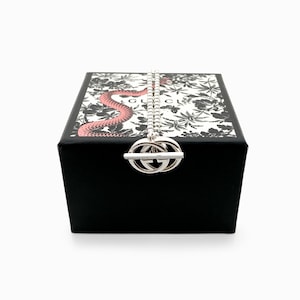 May include: A silver necklace with a double-G pendant rests on a black box. The box lid features a floral pattern with a pink snake design. The box is black with a white floral pattern and the word "GUCCI" is visible.
