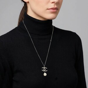 May include: Silver-toned necklace with a pendant featuring interlocking Cs encrusted with small crystals and a dangling white pearl. The necklace is worn against a black turtleneck.