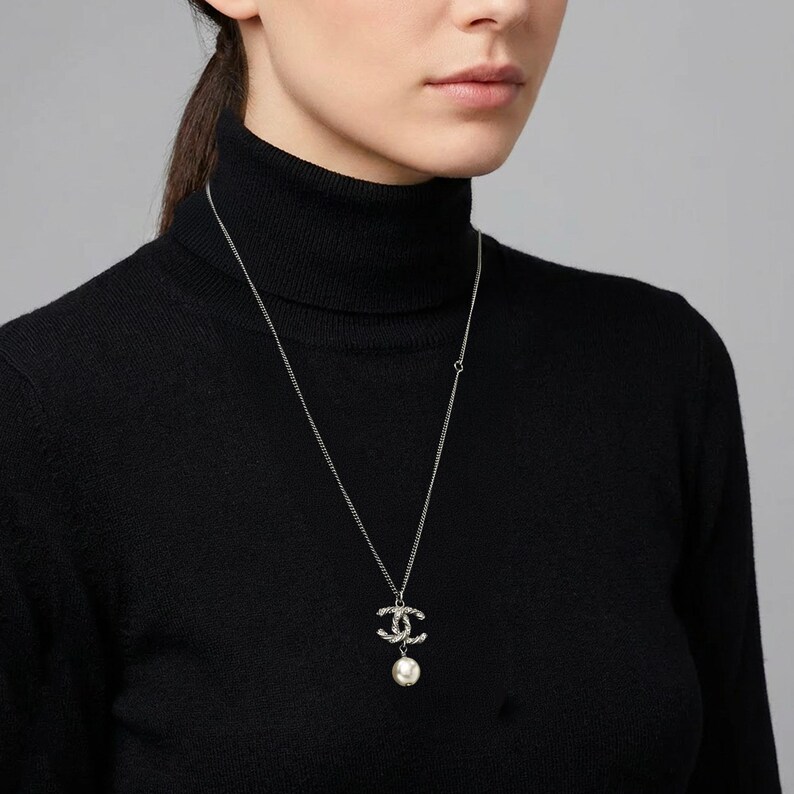 May include: Silver-toned necklace with a pendant featuring interlocking Cs encrusted with small crystals and a dangling white pearl. The necklace is worn against a black turtleneck.