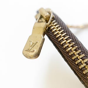 May include: Close-up of a brown and gold zipper with a gold zipper pull. The zipper pull features the embossed "LV" logo. The zipper is on a brown fabric with a repeating pattern.