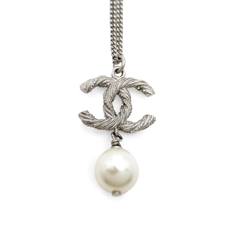 May include: Silver-tone necklace featuring a textured double-C pendant and a large, round, white pearl drop. The chain is a delicate, linked design. The pendant has a woven appearance. The pearl is smooth and lustrous.