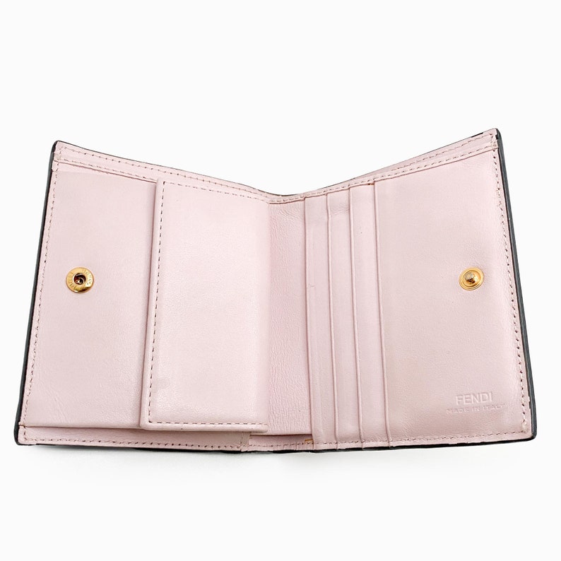 May include: A black leather wallet with a pink interior. The wallet has multiple card slots and a billfold. The wallet is open and the interior is visible. The wallet has a gold Fendi logo on the interior.