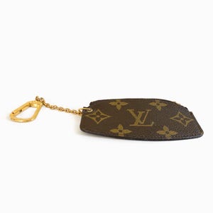 May include: A brown and tan key pouch with a gold-colored chain and clasp. The pouch features a repeating floral and logo pattern. The pouch is a small, rectangular shape with a curved edge.