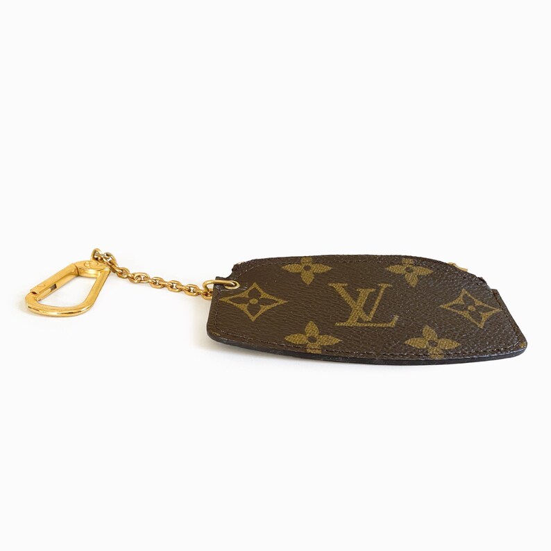 May include: A brown and tan key pouch with a gold-colored chain and clasp. The pouch features a repeating floral and logo pattern. The pouch is a small, rectangular shape with a curved edge.