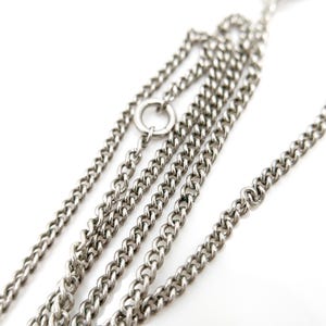 May include: Close-up of a silver-colored chain necklace with multiple strands. The chain links are small and closely connected, creating a textured surface. A small ring connects some of the chains. The background is white.