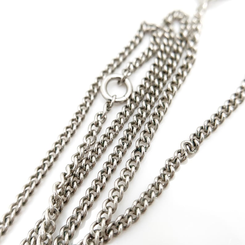 May include: Close-up of a silver-colored chain necklace with multiple strands. The chain links are small and closely connected, creating a textured surface. A small ring connects some of the chains. The background is white.