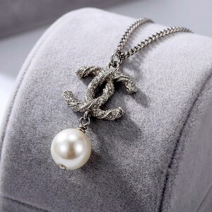 May include: Silver-toned necklace featuring a double-C pendant encrusted with small crystals and a dangling faux pearl. The chain is a delicate curb style. The necklace is displayed on a gray velvet surface.
