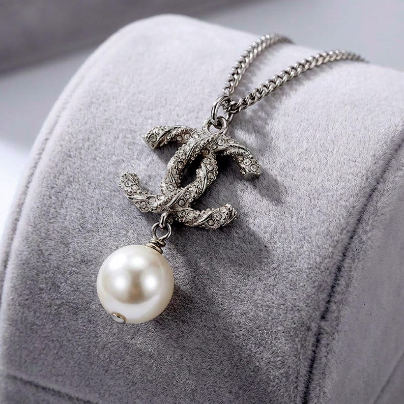 May include: Silver-toned necklace featuring a double-C pendant encrusted with small crystals and a dangling faux pearl. The chain is a delicate curb style. The necklace is displayed on a gray velvet surface.