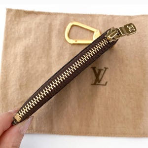May include: A brown zippered pouch with a gold-colored zipper and pull. A gold-colored carabiner clip is visible. The pouch is on a tan fabric with a gold-colored logo.