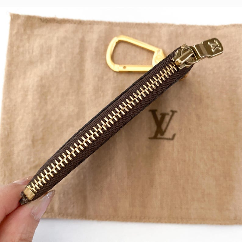 May include: A brown zippered pouch with a gold-colored zipper and pull. A gold-colored carabiner clip is visible. The pouch is on a tan fabric with a gold-colored logo.