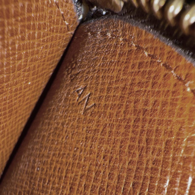 May include: Close-up of a brown leather surface with a textured pattern. The letters "AN" are embossed on the leather. The image shows a portion of a larger item, possibly a wallet or handbag.
