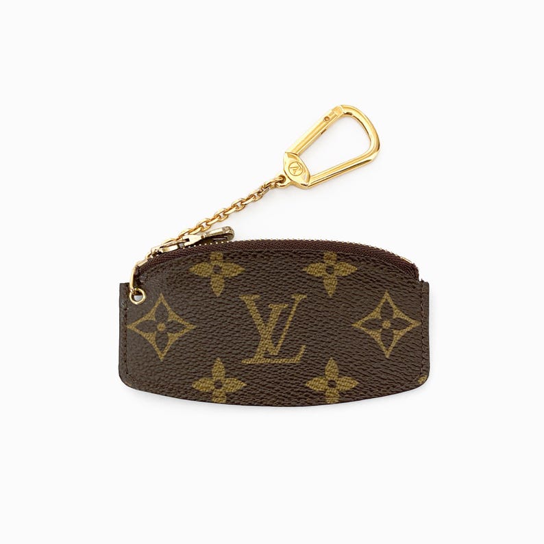 May include: Brown coin purse with a gold-tone zipper and key ring. The purse features a repeating pattern of brown floral shapes and the brand's logo in gold. The key ring is attached by a small gold chain.
