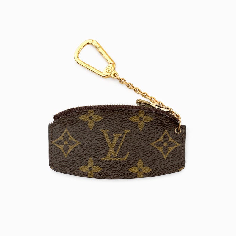 May include: Brown coin purse with a gold-colored chain and clasp. The purse features a repeating pattern of brown floral shapes and the letter "V" in a lighter brown color.