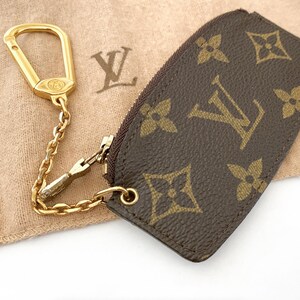 May include: A brown and tan key pouch with a gold-colored chain and clasp. The pouch features a repeating pattern of brown logos on a tan background. The clasp and chain are attached to the pouch with a small gold ring.
