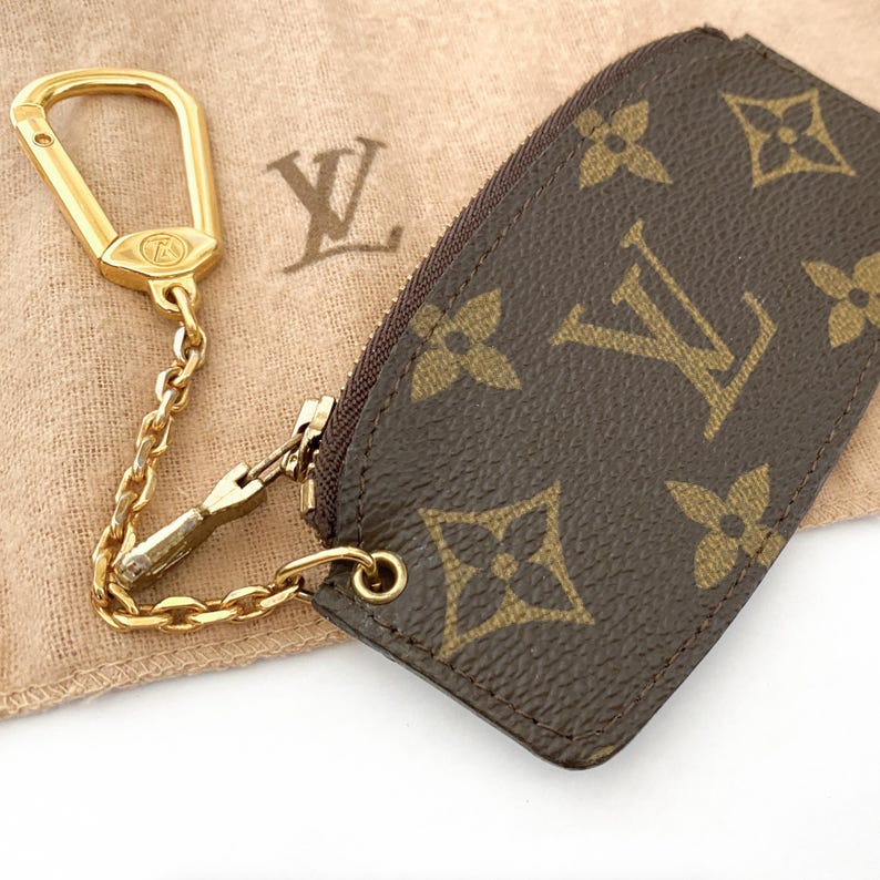 May include: A brown and tan key pouch with a gold-colored chain and clasp. The pouch features a repeating pattern of brown logos on a tan background. The clasp and chain are attached to the pouch with a small gold ring.