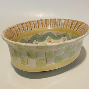 May include: An oval ceramic bowl featuring a hand-painted checkered pattern in shades of light green and white on the exterior. The rim is decorated with yellow and grey vertical stripes, while the interior displays a wavy teal line pattern.