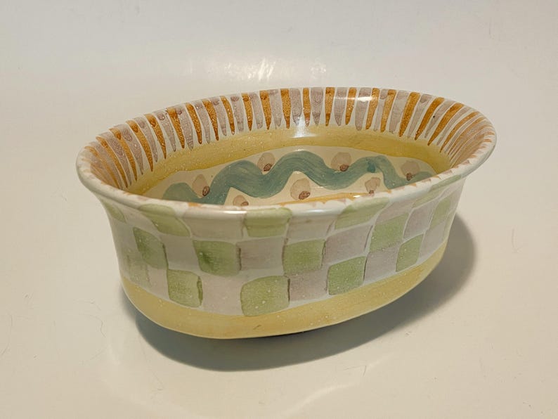 May include: An oval ceramic bowl featuring a hand-painted checkered pattern in shades of light green and white on the exterior. The rim is decorated with yellow and grey vertical stripes, while the interior displays a wavy teal line pattern.