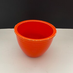 May include: A vibrant orange ceramic bowl with a rounded shape and a slightly distressed rim. The bowl has a smooth exterior and a bright red interior. The bowl is likely vintage and suitable for serving or display.