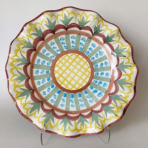 May include: A white ceramic plate with a scalloped edge and a colorful hand-painted design. The design features a central circle with a yellow and brown grid pattern, surrounded by concentric rings of blue, green, and red.