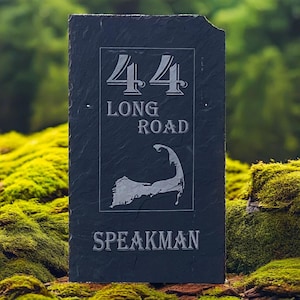 May include: A rectangular, dark gray slate house number sign. The sign features the number "44" above the words "LONG ROAD" and a coastal outline. The name "SPEAKMAN" is at the bottom. The sign is set against a backdrop of green moss.