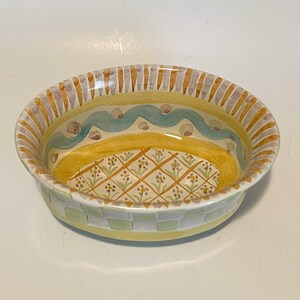 May include: An oval ceramic bowl featuring a yellow interior with a hand-painted grid pattern containing small floral motifs. The rim is decorated with alternating yellow and grey vertical stripes, while the exterior displays a light green checkerboard.