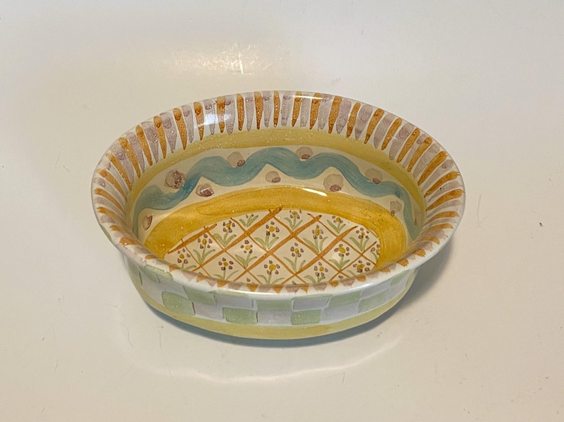 May include: An oval ceramic bowl featuring a yellow interior with a hand-painted grid pattern containing small floral motifs. The rim is decorated with alternating yellow and grey vertical stripes, while the exterior displays a light green checkerboard.